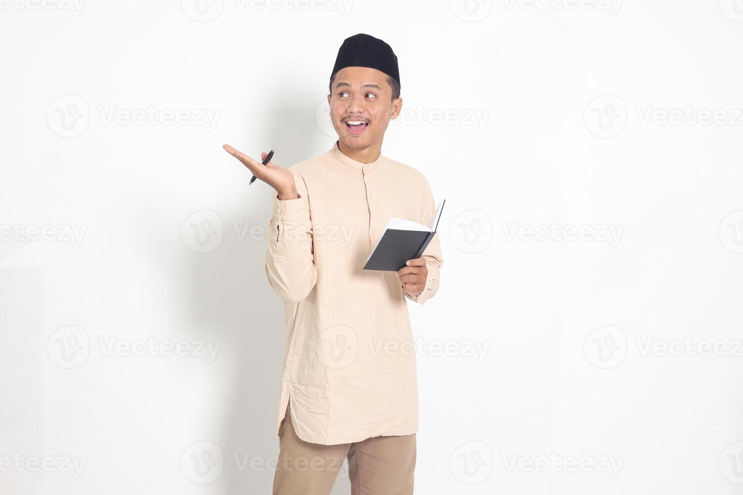 retrato do animado muçulmano homem dentro Koko camisa com peci lendo uma caderno. feliz ásia cara apontando para a lado com dedo. isolado imagem em branco fundo foto