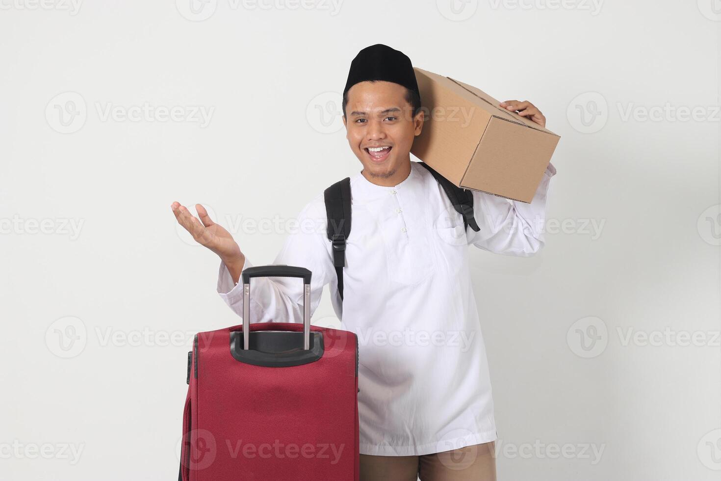 retrato do animado ásia muçulmano homem carregando cartão caixa e mala de viagem enquanto apontando lateralmente com dedo. indo casa para eid mubarak. isolado imagem em branco fundo foto