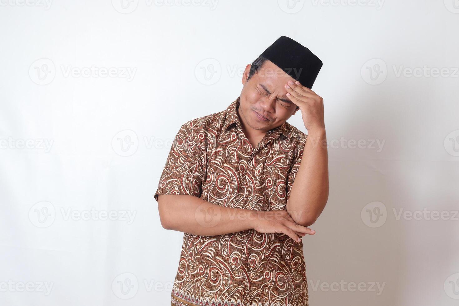 retrato do sofrimento ásia homem vestindo batik camisa e Songkok tendo dor de cabeça enquanto tocante dele testa área. isolado imagem em cinzento fundo foto