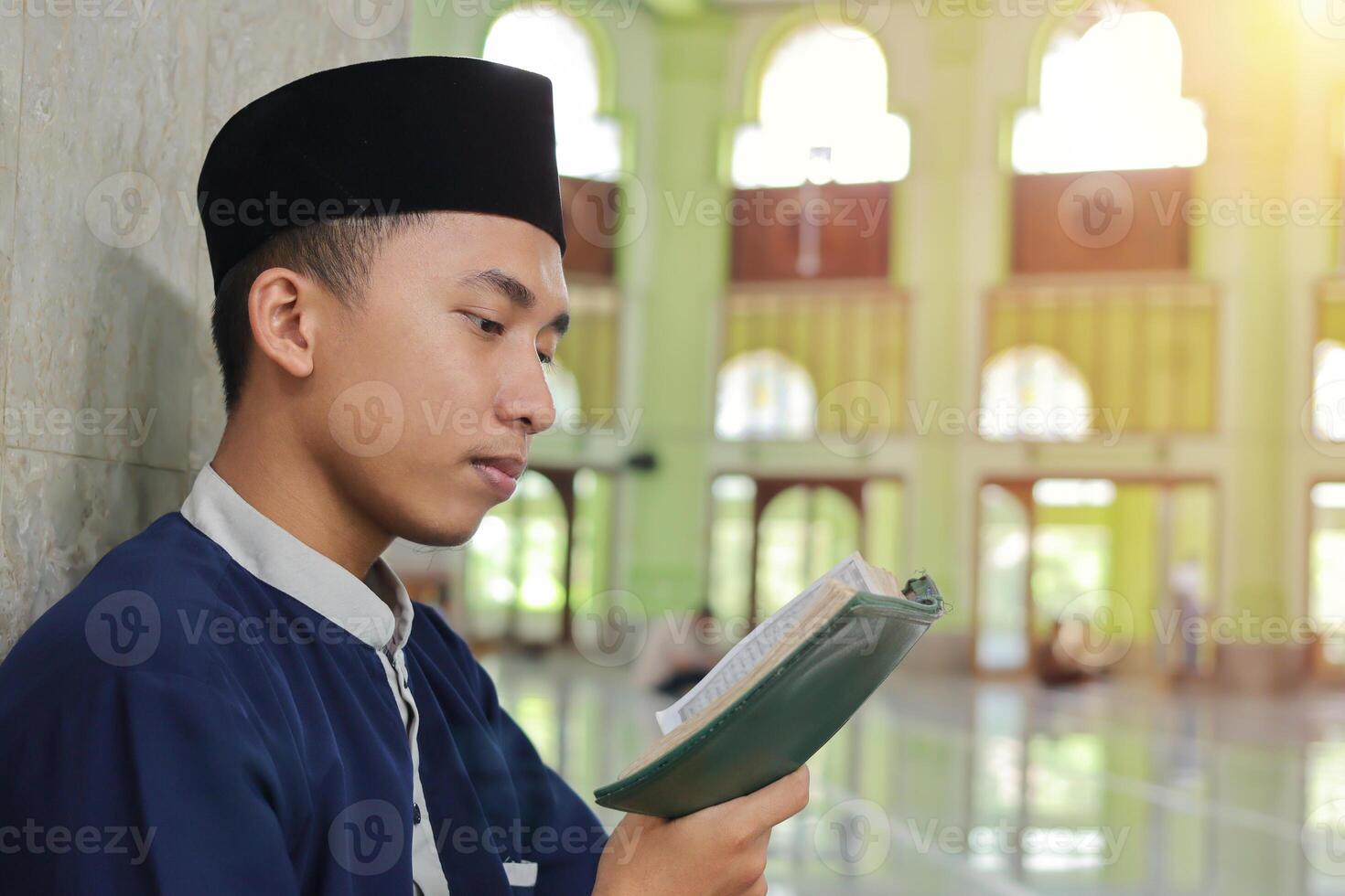 religioso ásia homem dentro muçulmano camisa e Preto boné lendo a piedosos livro do Alcorão dentro a público mesquita foto