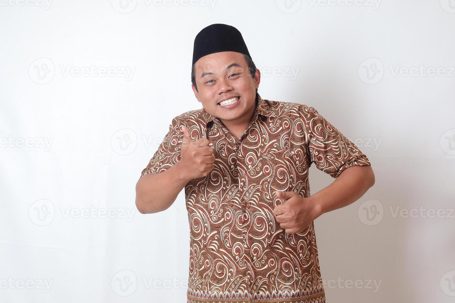 retrato do animado ásia homem vestindo batik camisa e Songkok sorridente e olhando às Câmera, fazer polegares acima mão gesto. isolado imagem em cinzento fundo foto