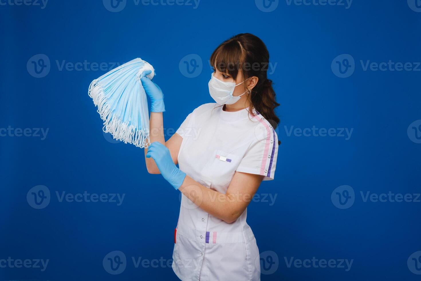 uma jovem menina médico dentro uma médico mascarar detém uma muitos do máscaras dentro dela mãos. uma enfermeira poses em uma azul fundo foto