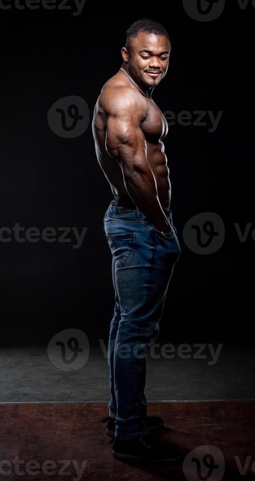 muito muscular afro americano homem com nu tronco poses em Preto fundo. fisiculturista dentro jeans olhando às a Câmera foto