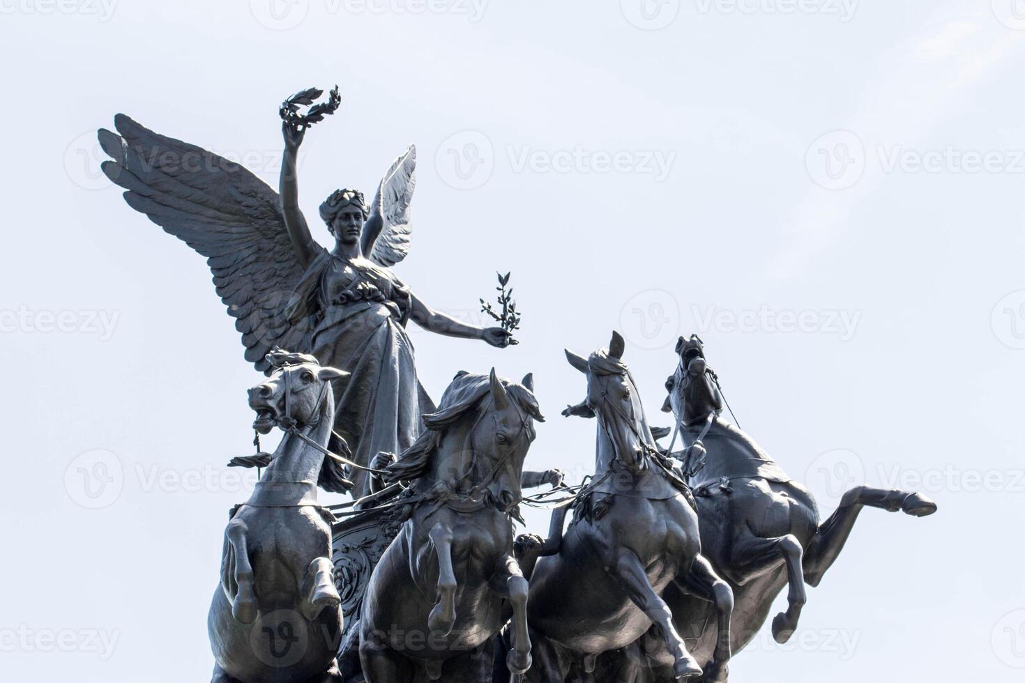 bronze escultura do uma alado fêmea figura com uma louro guirlanda, conduzindo uma carruagem puxado de quatro cavalos contra uma Claro céu. foto