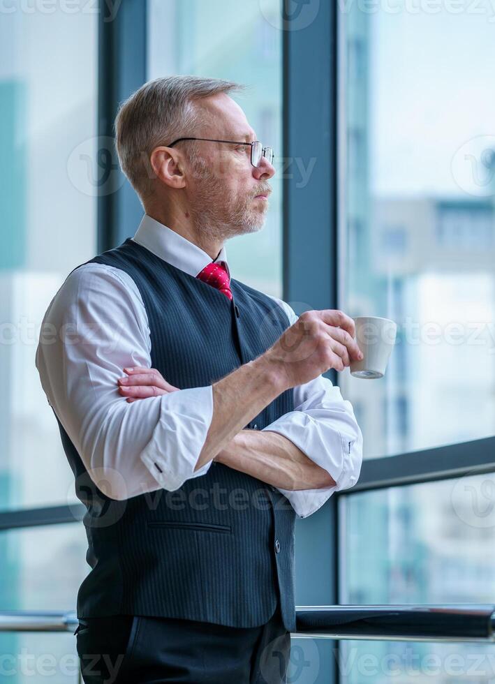 Senior homem de negocios desgasta branco camisa. bebendo café perto grande janela com cidade Visão . foto a partir de a lado.