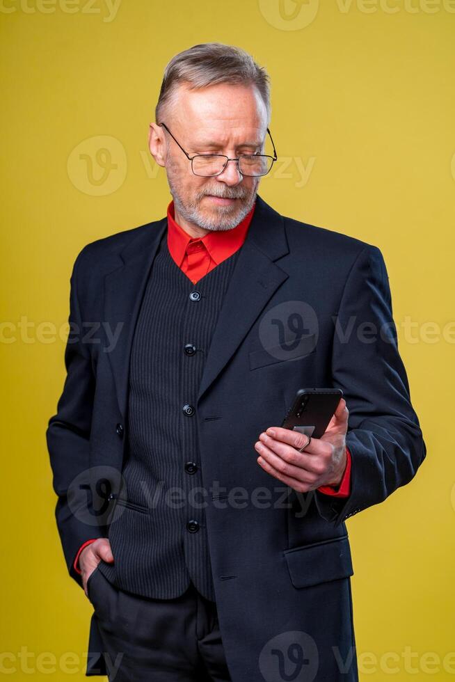 retrato do Senior homem de negocios segurando dentro mãos telefone enquanto em pé às estúdio sobre amarelo fundo. o negócio pessoas conceito. foto