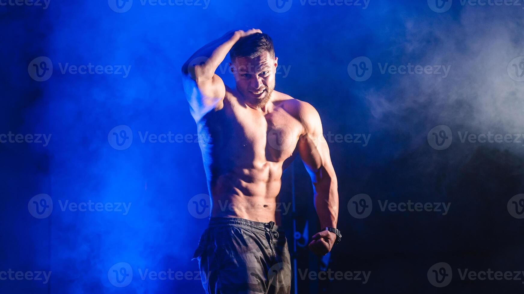 fisiculturista mostrando barriga e bíceps músculos. Forte homem flexão músculos. ginástica modelo posando às Câmera. Preto e azul luz fundo. atleta olhando em linha reta às Câmera foto