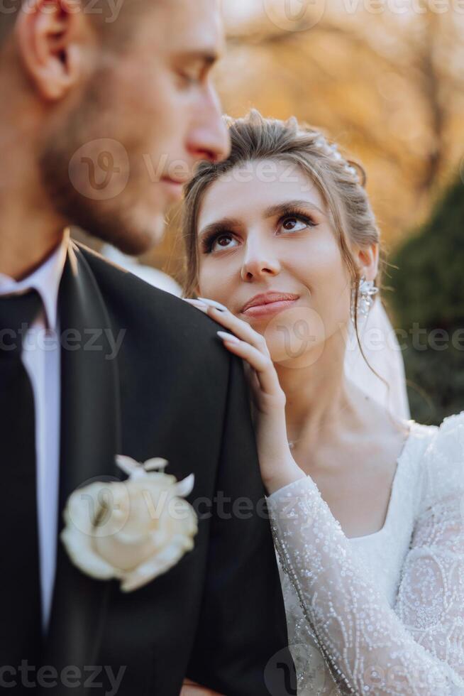 adorável e à moda recém-casados estão abraçando e sorridente contra a fundo do outono natureza dentro uma lindo jardim. a incrivelmente lindo jovem noiva inclinou-se contra a ombro do dela Amado noivo foto