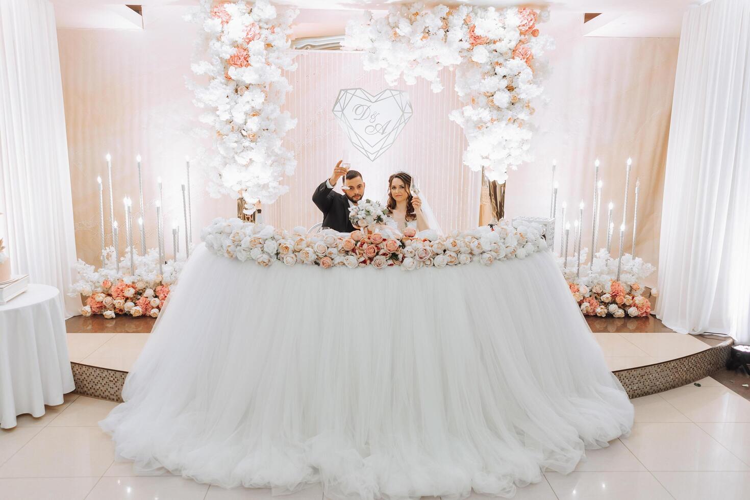 uma jovem noivo dentro uma Preto terno e uma sorridente morena noiva estão às seus mesa dentro uma banquete salão, levantando uma torrada contra uma fundo do fresco flores foto