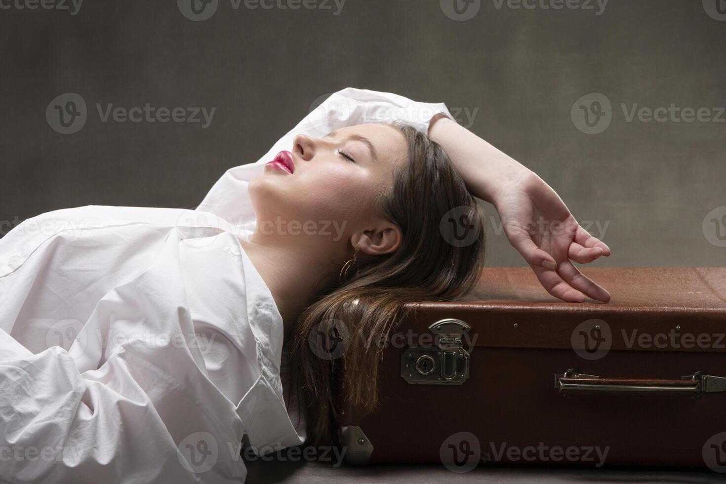 uma lindo menina mentiras em a velho mala de viagem em uma cinzento fundo. foto