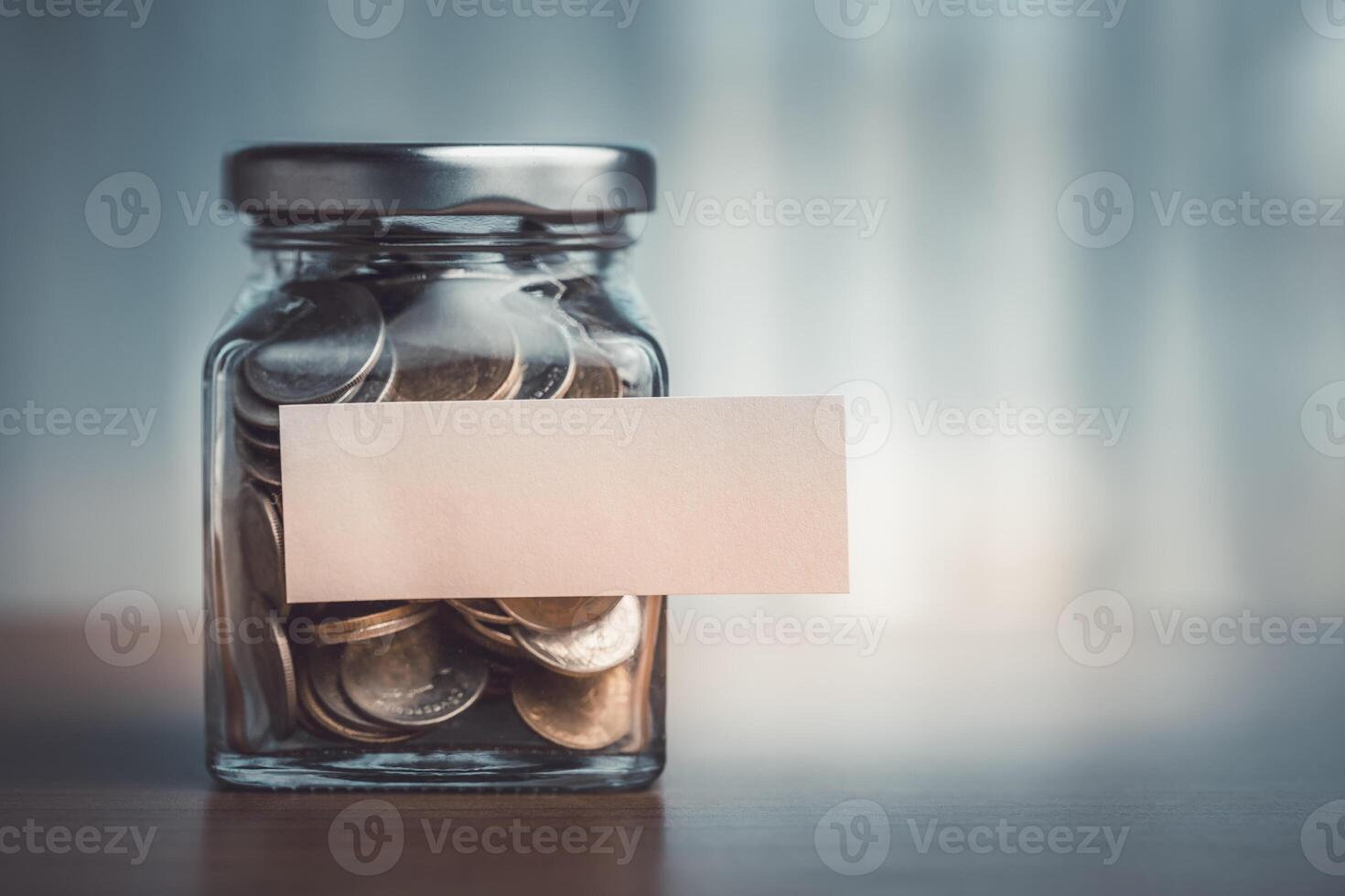 moedas dentro uma vidro jarra com em branco papel rótulo em de madeira mesa foto