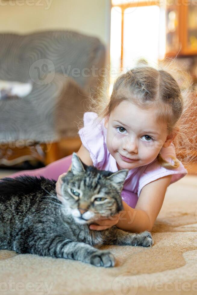 pouco menina com cinzento gato deitado em tapete dentro casa vivo quarto foto