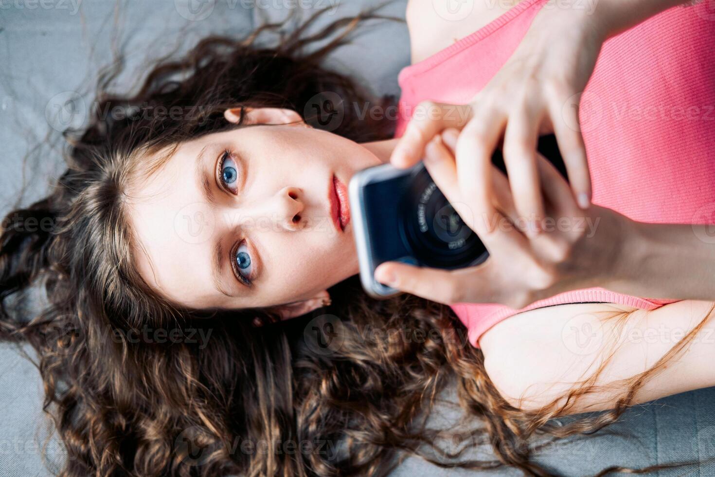 jovem mulher com grandes encaracolado cabelo mentiras em a sofá e pergaminhos social redes em dela telefone dentro uma Rosa principal, topo Visão foto