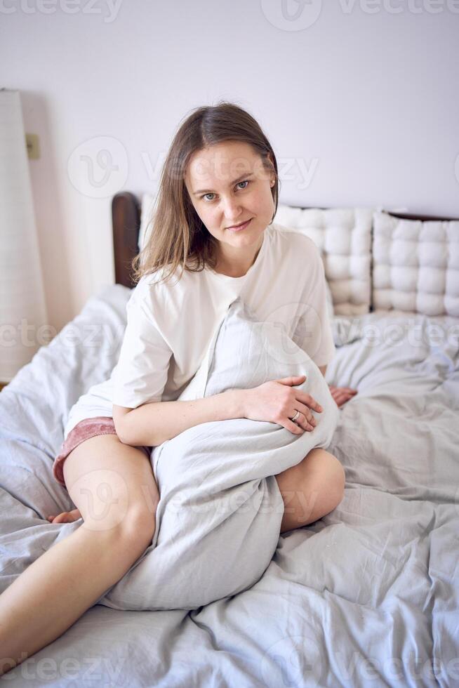 lindo jovem mulher dentro branco camiseta em cama abraçando travesseiro foto
