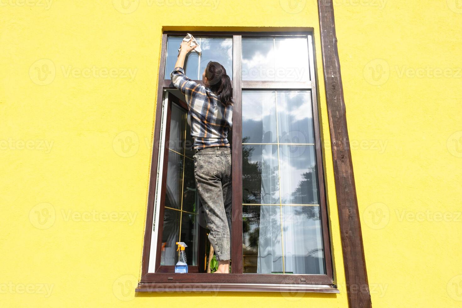 uma mulher manualmente lavagens a janela do a casa com uma trapo com uma spray limpador e uma esfregão fora. segurança às altura, restaurando ordem e limpeza dentro a primavera, limpeza serviço foto