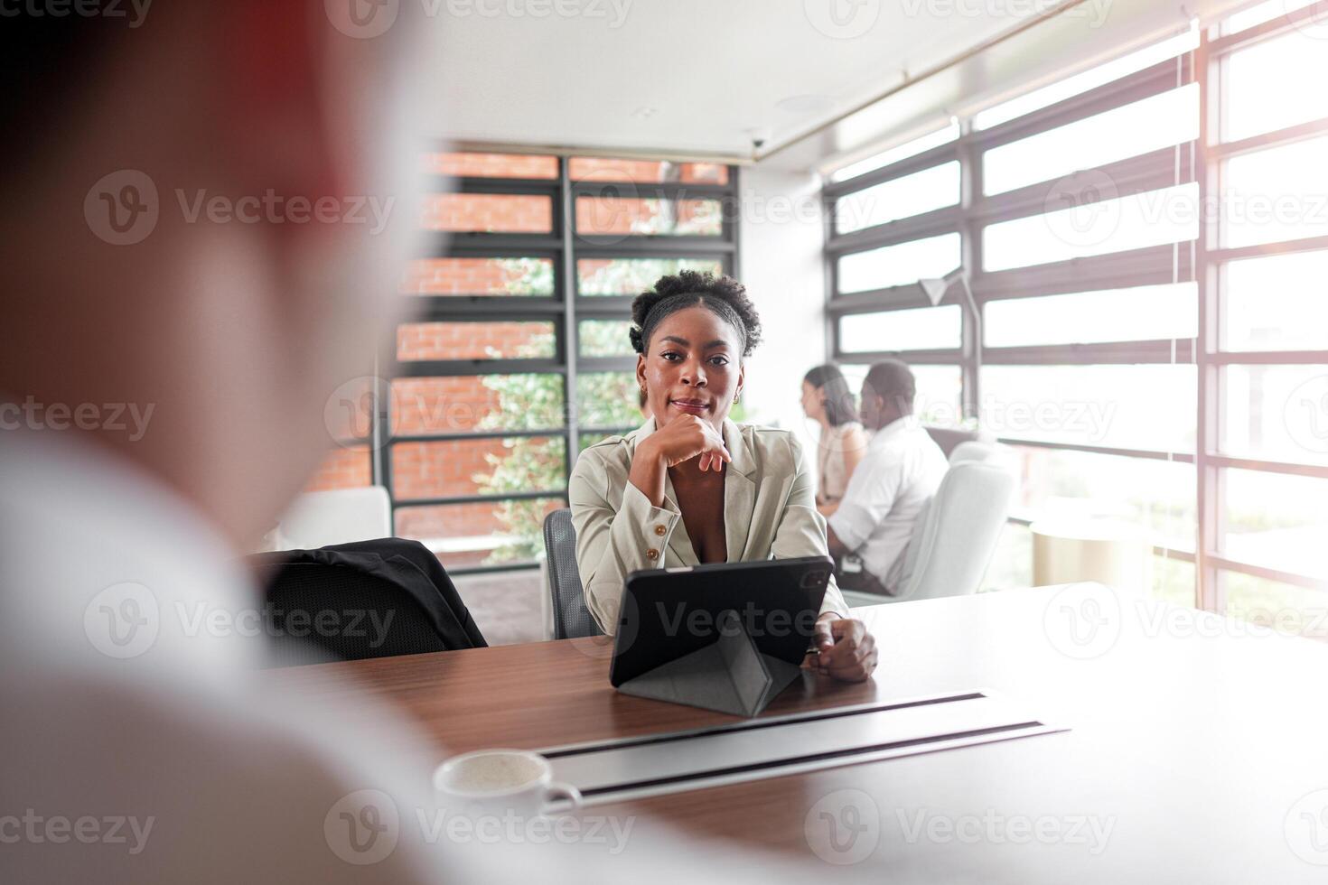 atraente africano jovem confiante empresária sentado às a escritório mesa com grupo do colegas dentro a fundo. trabalhando em computador portátil computador. bem sucedido mulher empreendedor. entrevista Novo funcionários. foto