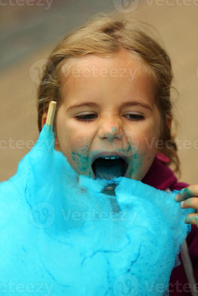 fechar-se retrato do feliz criança pequena menina comendo brilhante azul algodão doce foto