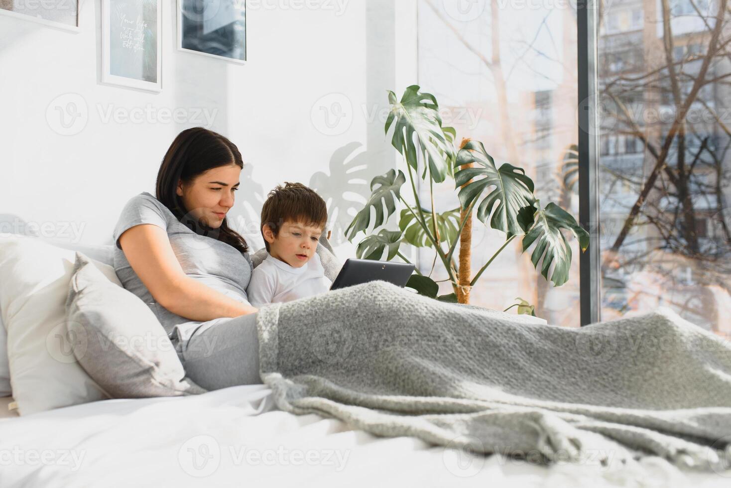 lindo sorridente mãe e filho jogando com digital tábua deitado em cama. foto