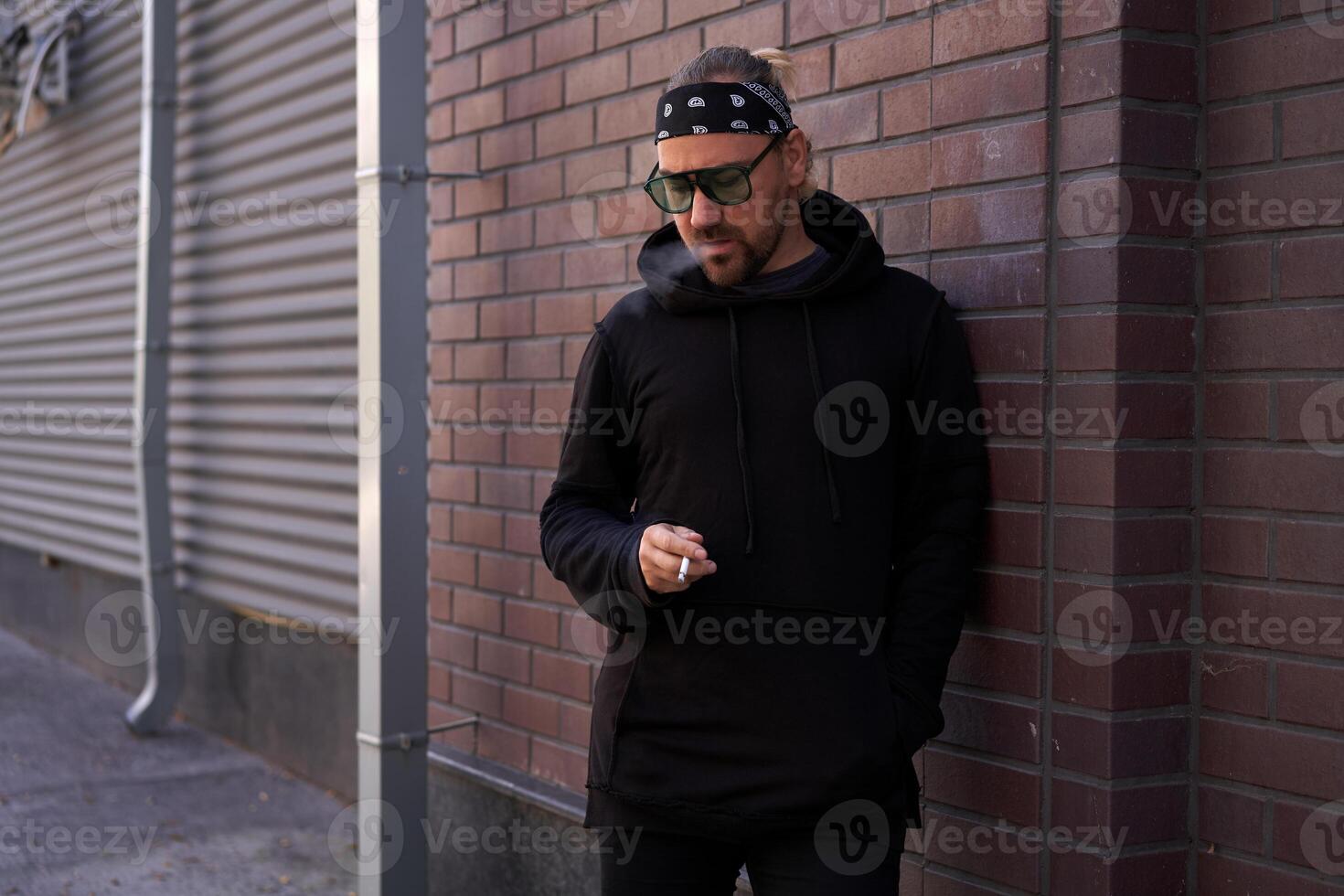 bonito jovem com a barba por fazer homem Preto moletom com capuz e bandana carrinhos quintal perto tijolo paredes verão dia. foto