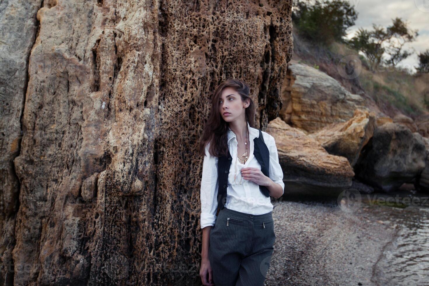 o negócio mulher em terno colete e branco camisa em pé perto grande pedra em a mar custo foto