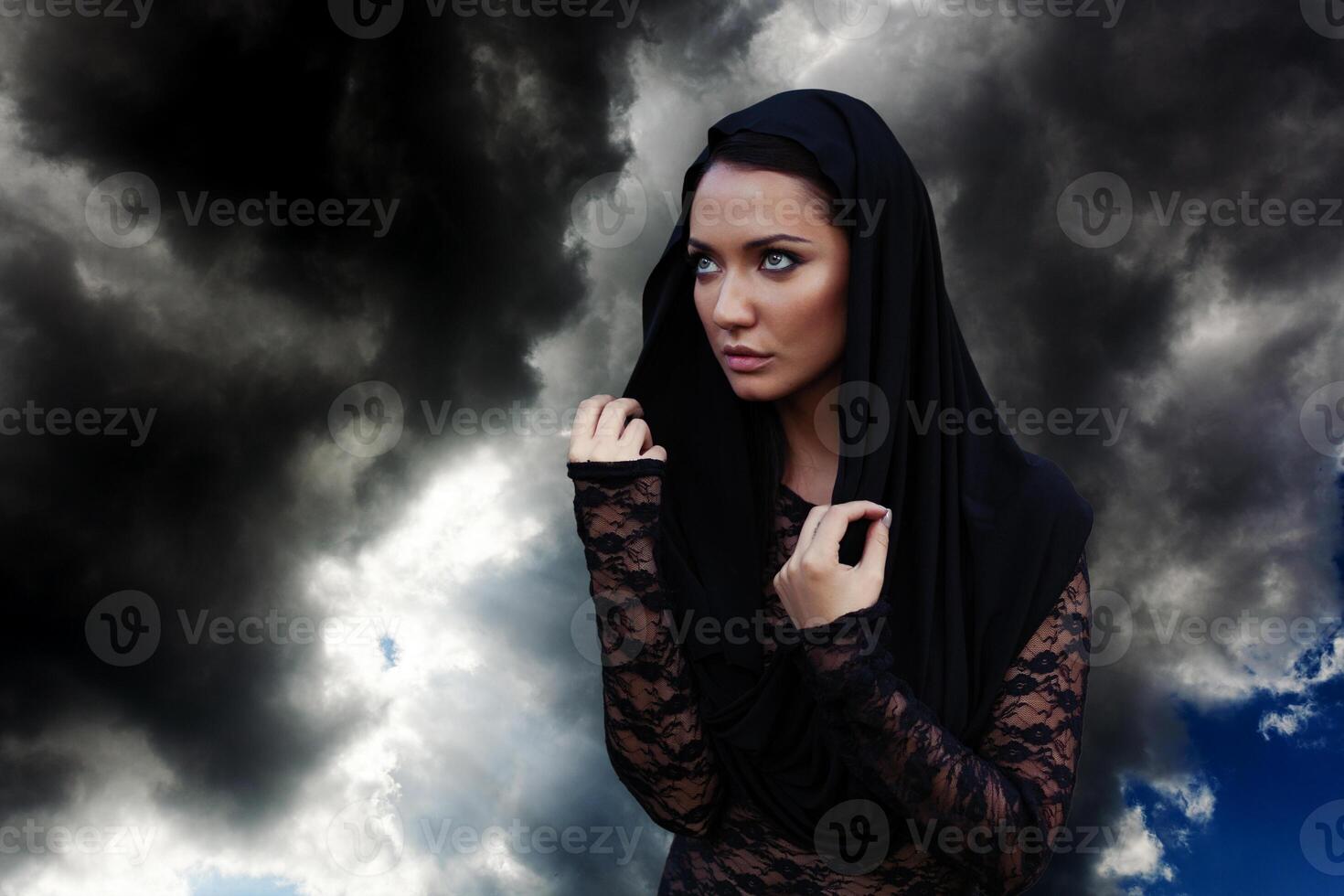 jovem lindo mulher com a Preto cabelo e dentro a Preto de capuz e Preto transparente blusa às a céu fundo foto