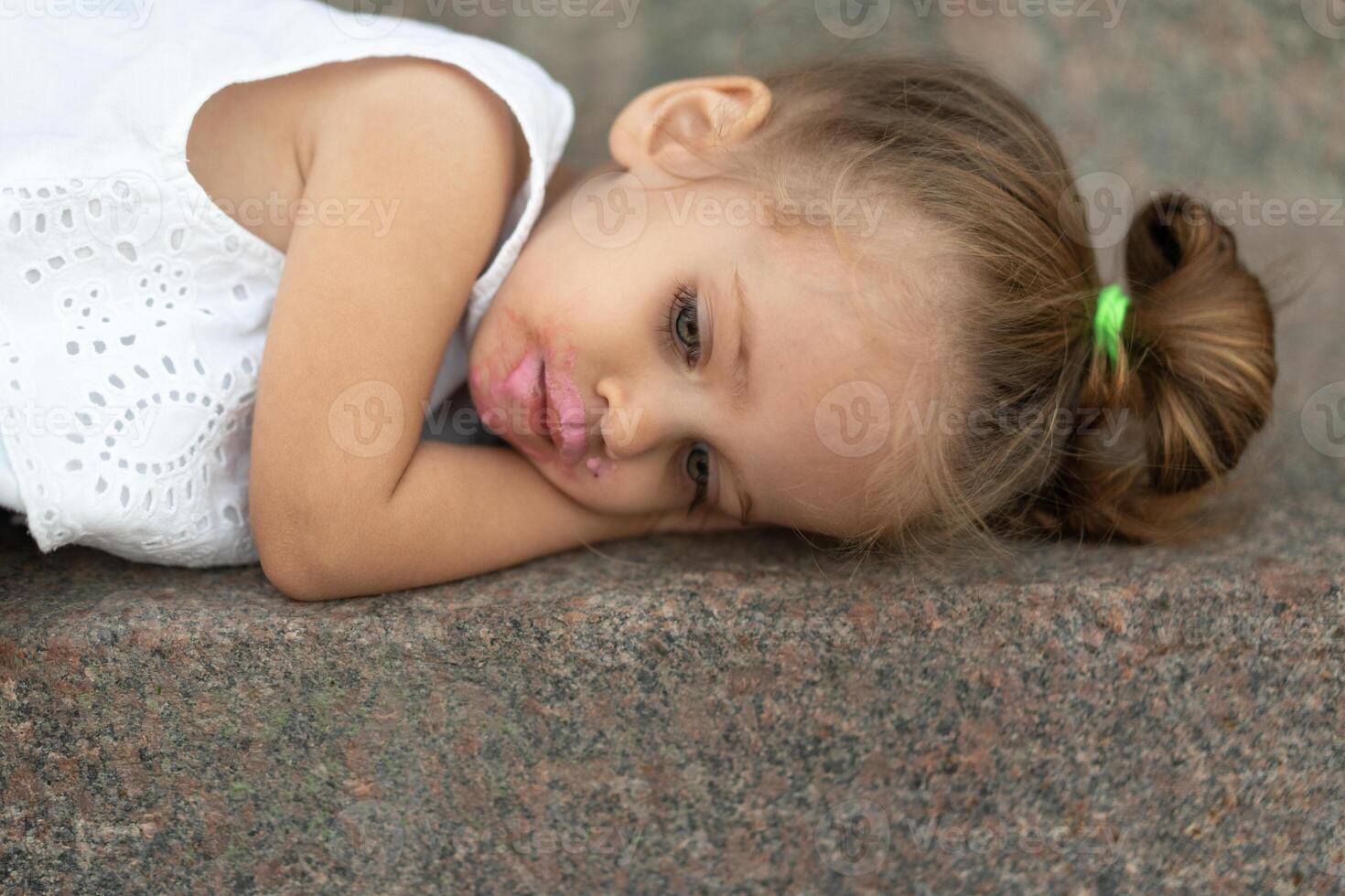 uma lindo pequeno menina do europeu aparência Loiras cabelo deitado triste em rua asfalto. foto