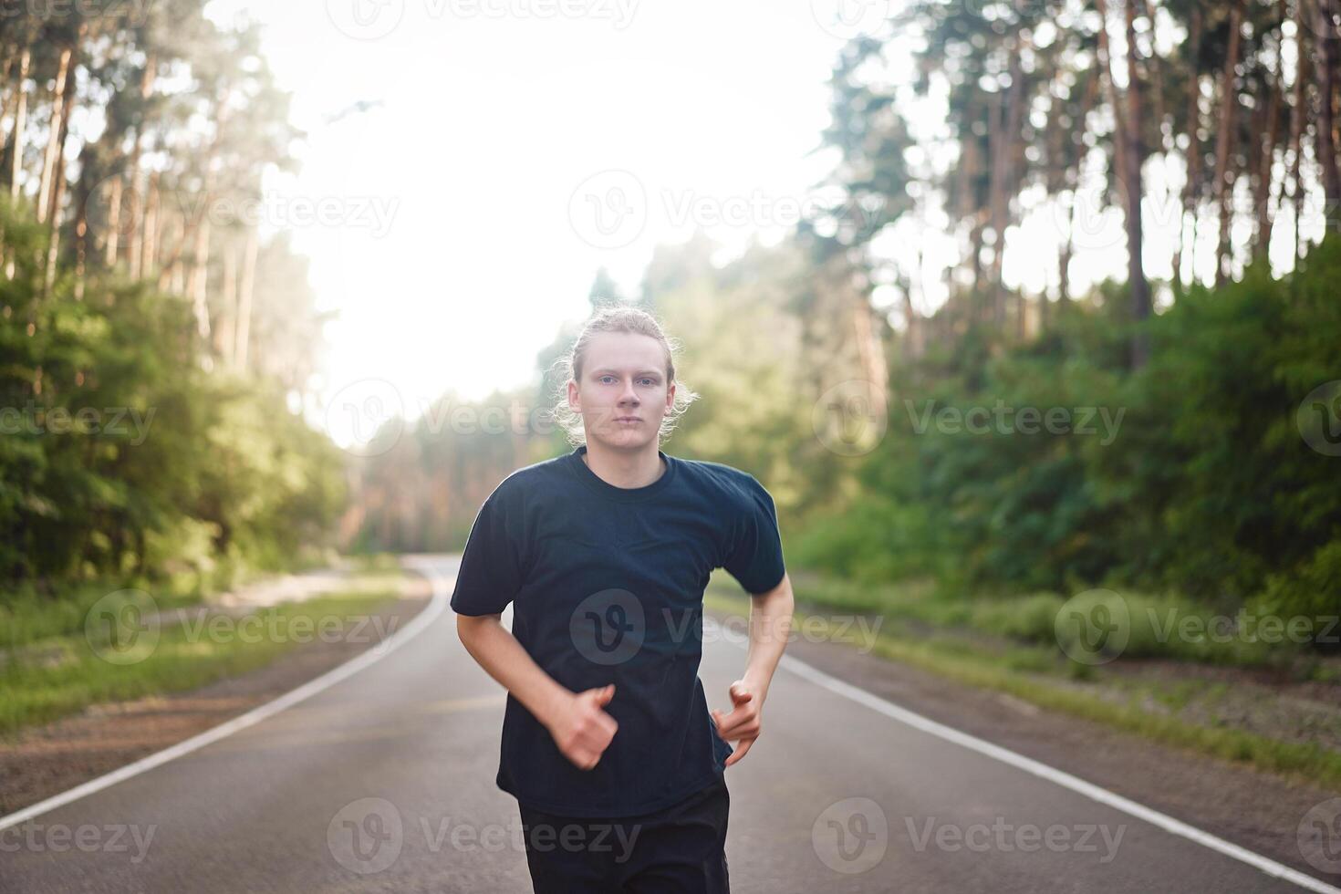 caucasiano jovem encaracolado homem atleta corre ensolarado verão dia em asfalto estrada dentro a floresta. foto
