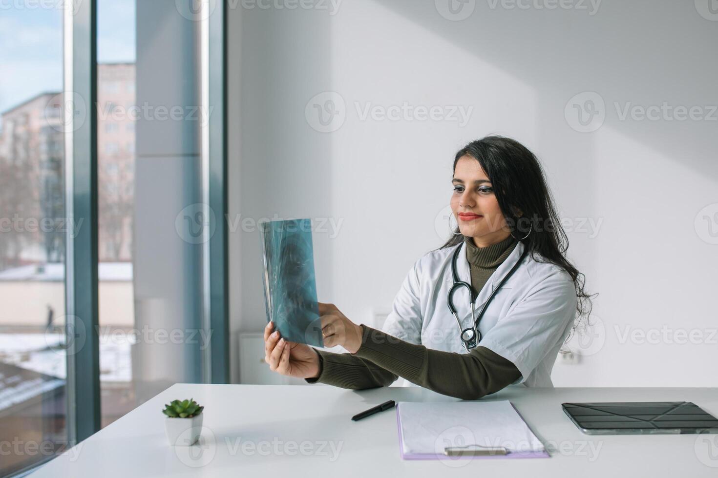 indiano fêmea médico dentro branco casaco com estetoscópio examina raio X e faz notas. jovem indiano médico aluna às a clínica dentro prática foto