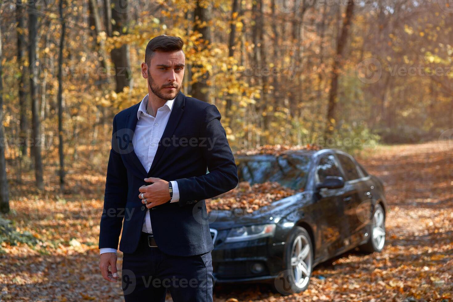 uma bonito jovem caucasiano homem vestindo o negócio terno em pé perto Preto carro em natureza. foto