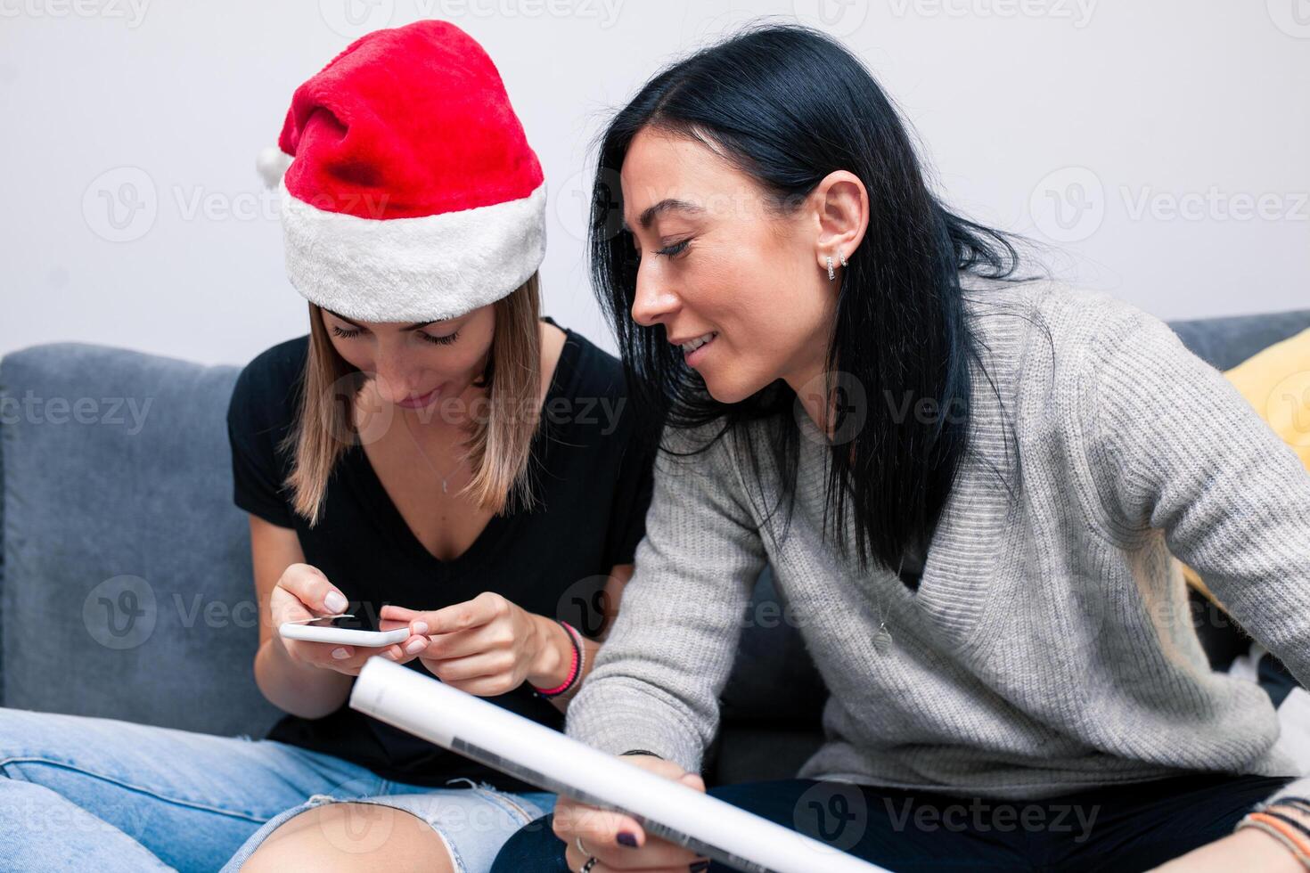 dois jovem mulheres estão preparando para a Novo ano olhando dentro Móvel telefone maneiras para pacote presentes. Novo ano celebração foto