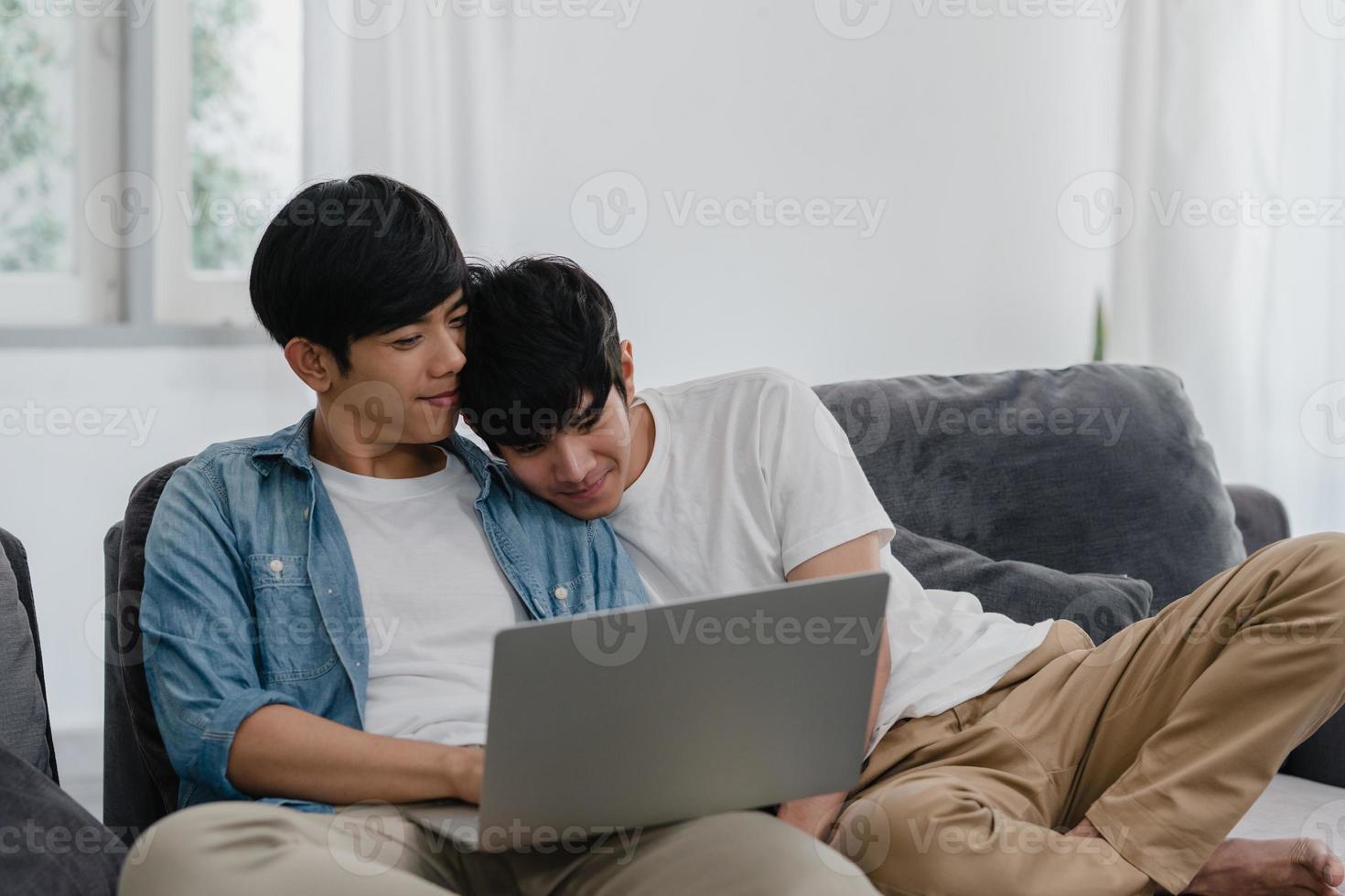 jovem casal gay usando computador portátil em casa moderna. homens lgbtq asiáticos felizes relaxam a diversão usando tecnologia, assistindo filme na internet juntos, enquanto estava deitado sofá na sala de estar no conceito de casa. foto