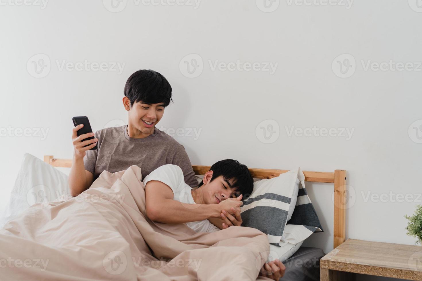 casal gay asiático usando telefone celular em casa. jovem ásia lgbtq homem feliz relaxar descansar depois de acordar, verificar a mídia social enquanto seu namorado dorme deitado na cama no quarto em casa no conceito de manhã. foto