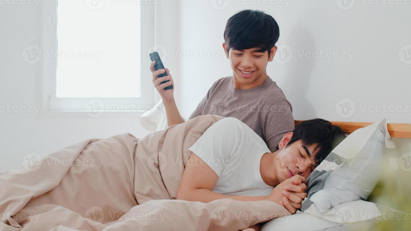 casal gay asiático usando telefone celular em casa. jovem ásia lgbtq homem feliz relaxar descansar depois de acordar, verificar a mídia social enquanto seu namorado dorme deitado na cama no quarto em casa no conceito de manhã. foto