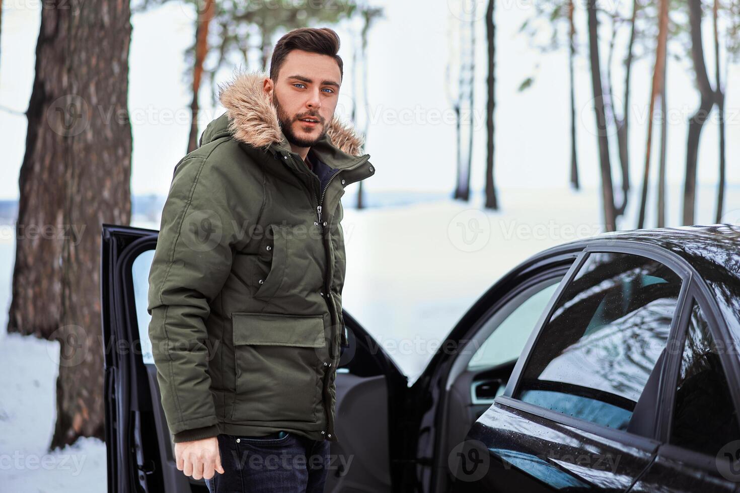 jovem adulto bonito homem em pé dentro inverno floresta perto dele carro. atraente caucasiano cara esperando para Socorro quando carro é quebrado. foto