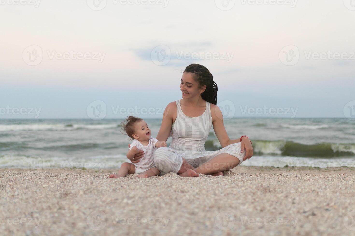 feliz família mãe e criança filha fazendo ioga, meditar dentro lótus posição em de praia às pôr do sol foto