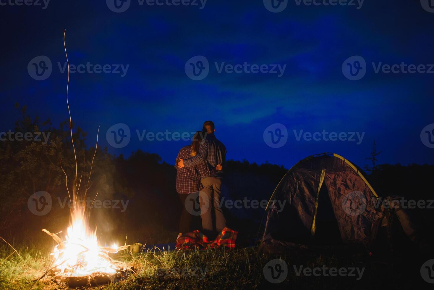 amoroso casal caminhantes desfrutando cada outro, em pé de fogueira às noite debaixo tarde céu perto árvores e barraca. romântico acampamento perto floresta dentro a montanhas foto