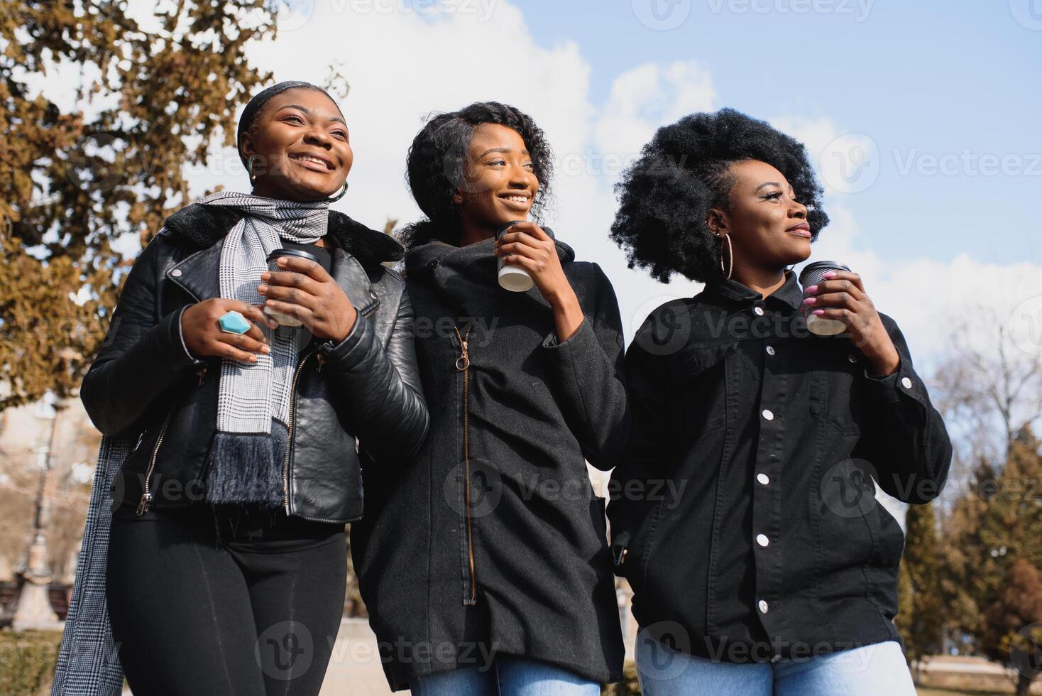 três lindo e à moda pele escura meninas com grandes cabelo em pé dentro uma cidade e bebendo uma café e usar a telefones foto