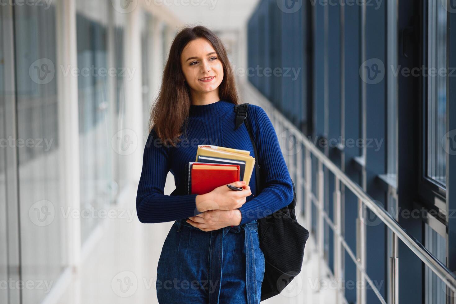 alegre morena aluna menina com Preto mochila detém livros dentro moderno prédio. fêmea aluna em pé com livros dentro Faculdade corredor foto