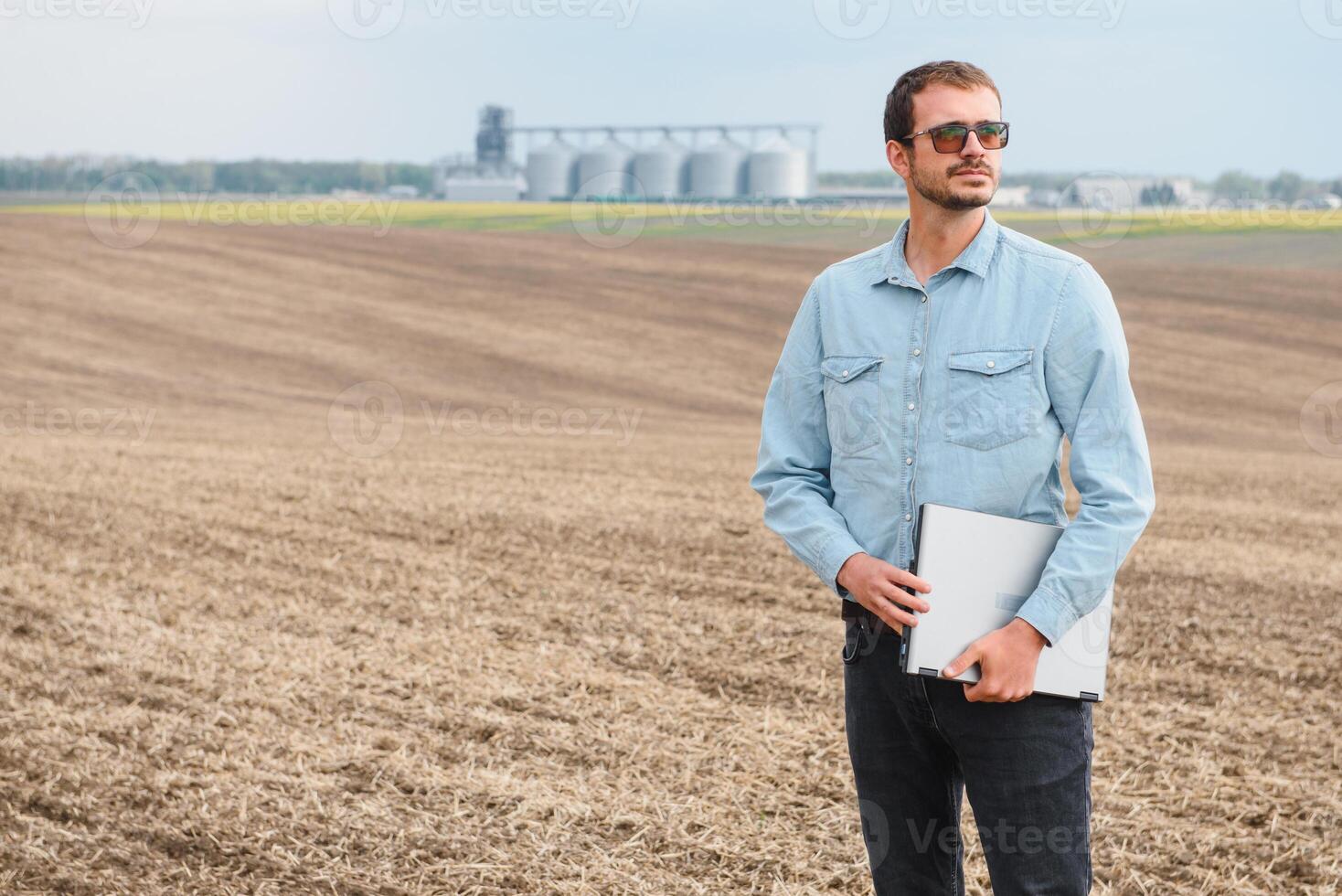 feliz agricultor com computador portátil em pé dentro trigo campo dentro frente do grão silo foto