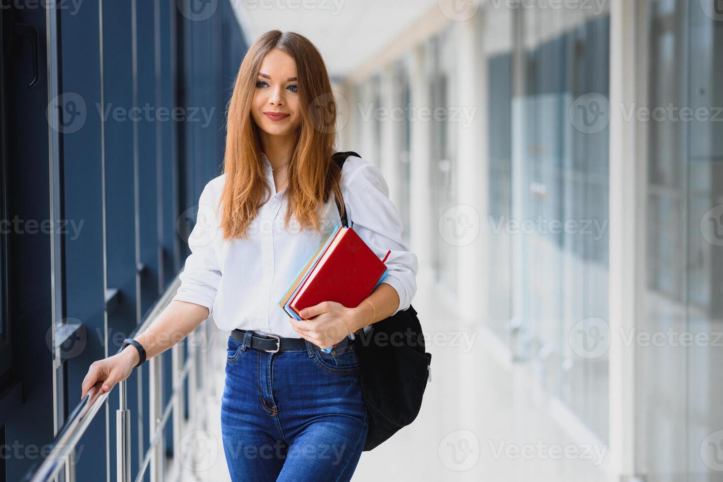 positividade lindo menina sorridente às Câmera, em pé em corredor com notas Como mochila, indo para lição. feliz morena fêmea aluna estudando dentro luxo universidade. foto