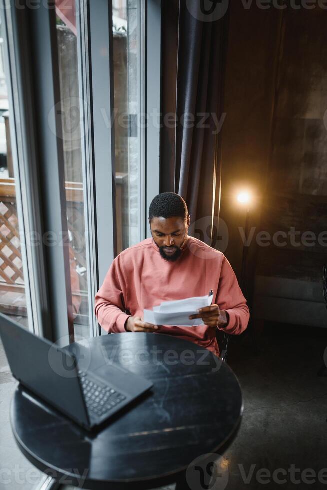 retrato do feliz africano homem de negocios sentado dentro uma cafeteria e trabalhando em computador portátil. foto