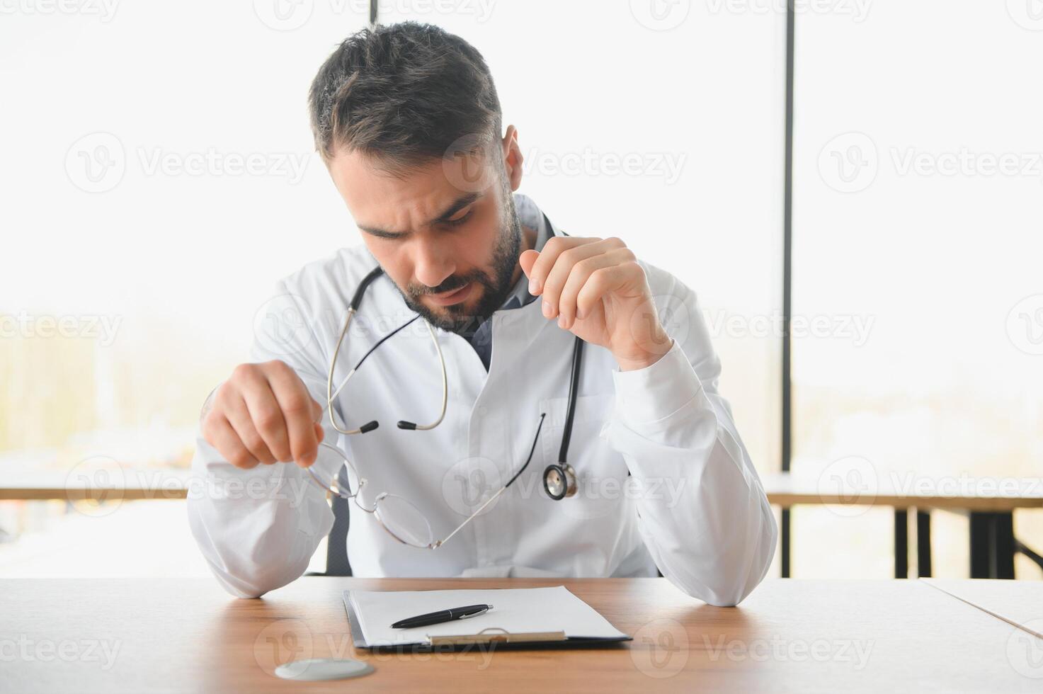 bonito médico homem vestindo médico uniforme sentado em dele local de trabalho cansado segurando dele cabeça sentindo-me fadiga e dor de cabeça. estresse e frustração conceito foto