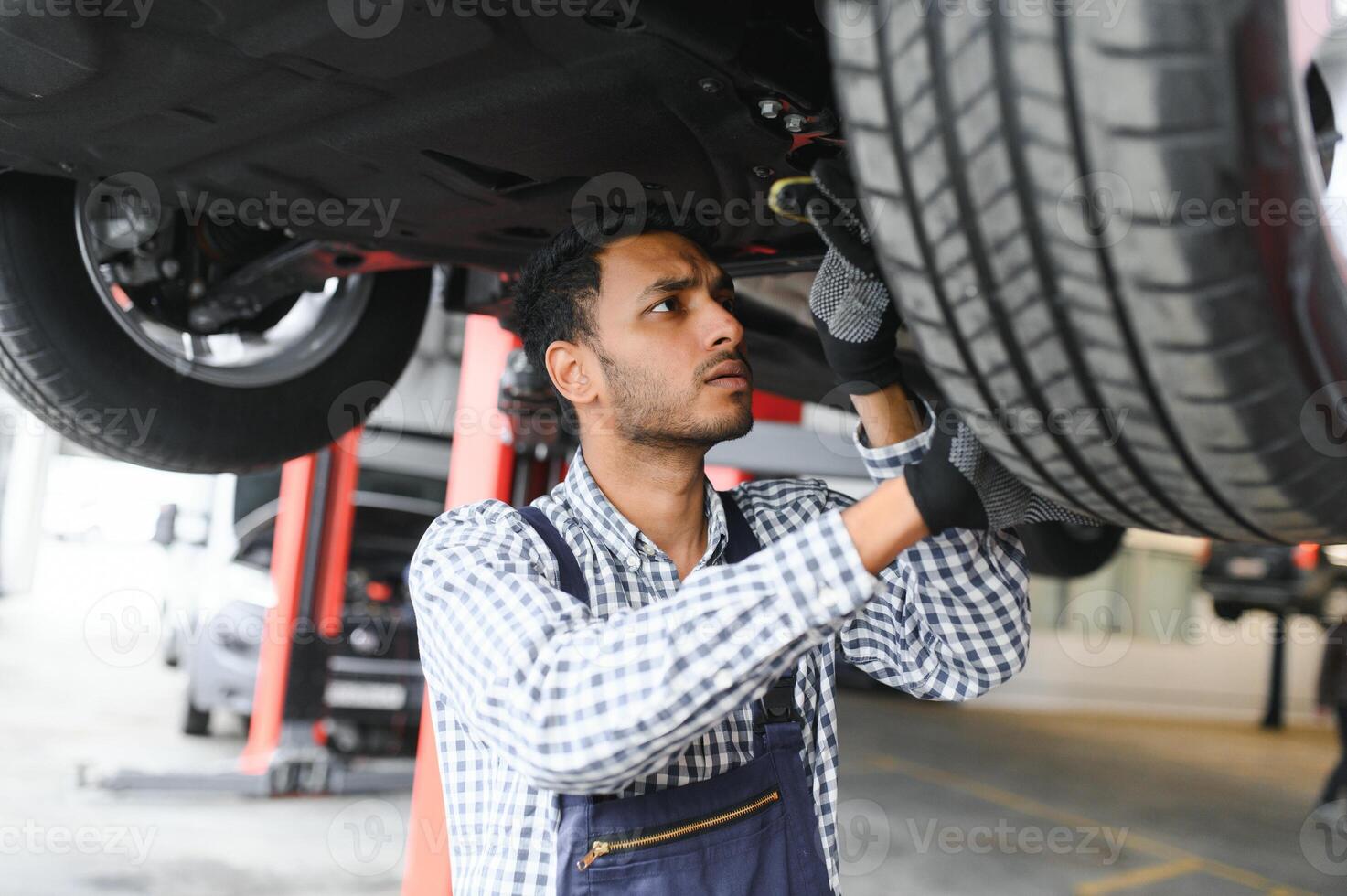 indiano carro mecânico em pé e trabalhando dentro serviço estação. carro especialistas examinando a levantado carro. profissional reparadores vestindo mecânico uniforme dentro azul cor. foto