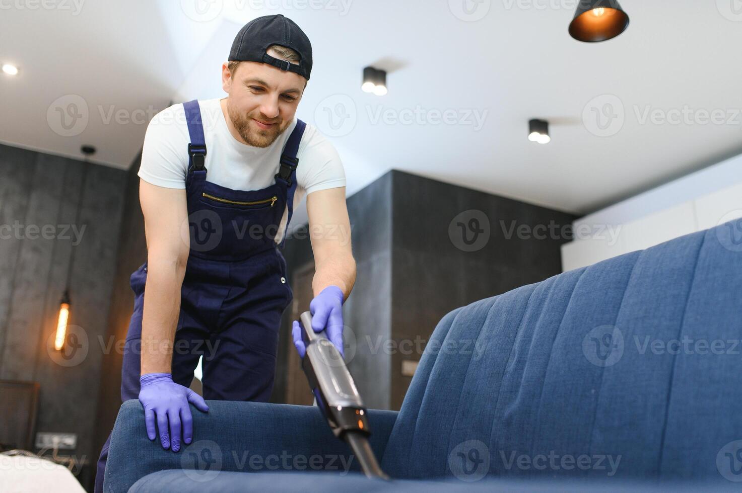 limpeza serviço. homem zelador dentro luvas e uniforme vácuo limpar \ limpo sofá com profissional equipamento. foto