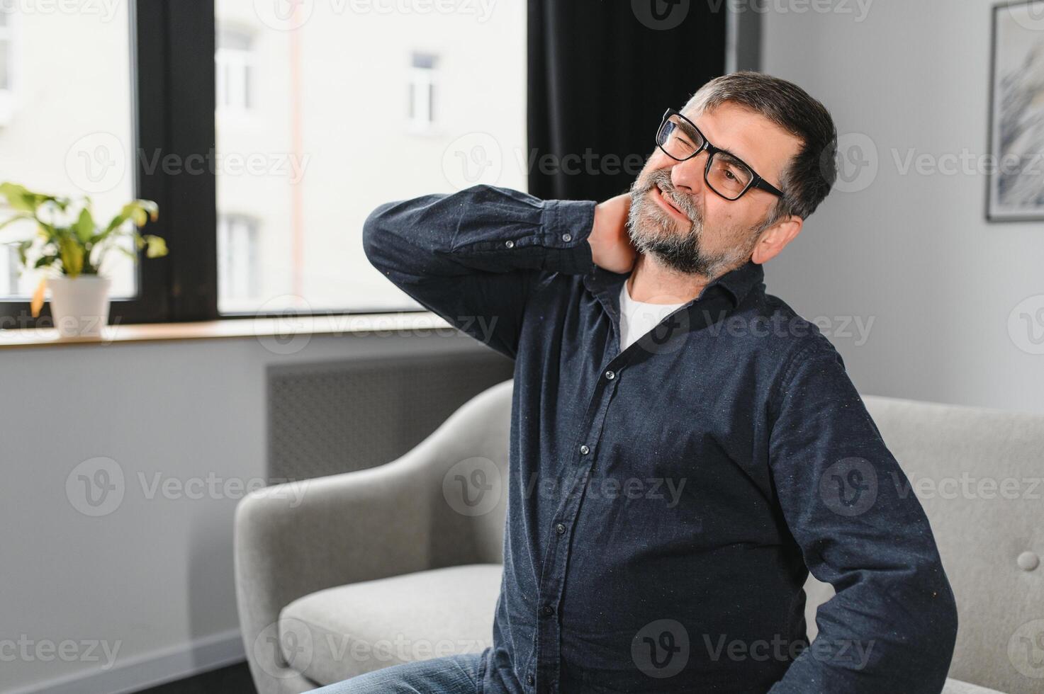 pescoço dor. retrato do Exausta maduro homem massageando dolorido pescoço sofrimento a partir de dor sentado em sofá às casa dentro vivo sala. osteoartrite, Mais velho pessoas saúde problema, sedentário estilo de vida conceito foto