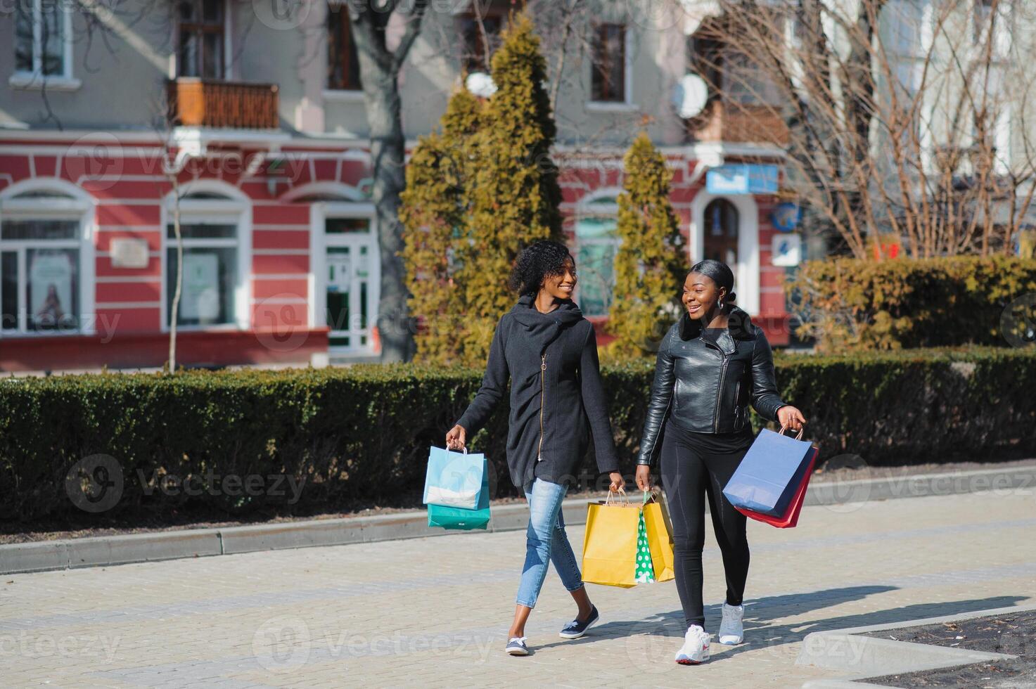 dois afro americano mulheres amigos dentro a cidade em uma compras viagem carregando colorida compras bolsas. foto