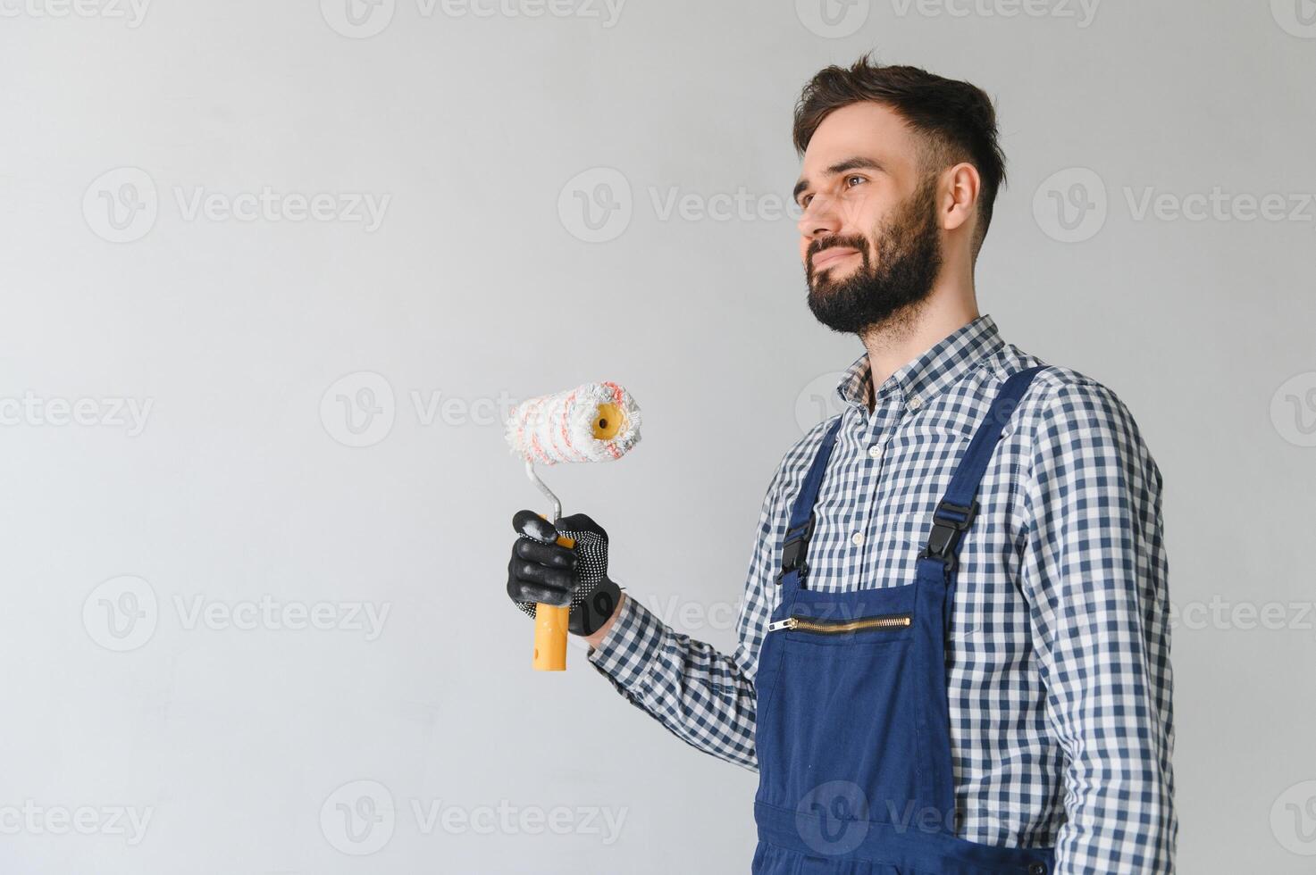 jovem sorridente profissional trabalhador dentro azul uniforme em pé com pintura rolo dentro Novo apartamento para reparação sobre cinzento paredes fundo, cópia de espaço. foto