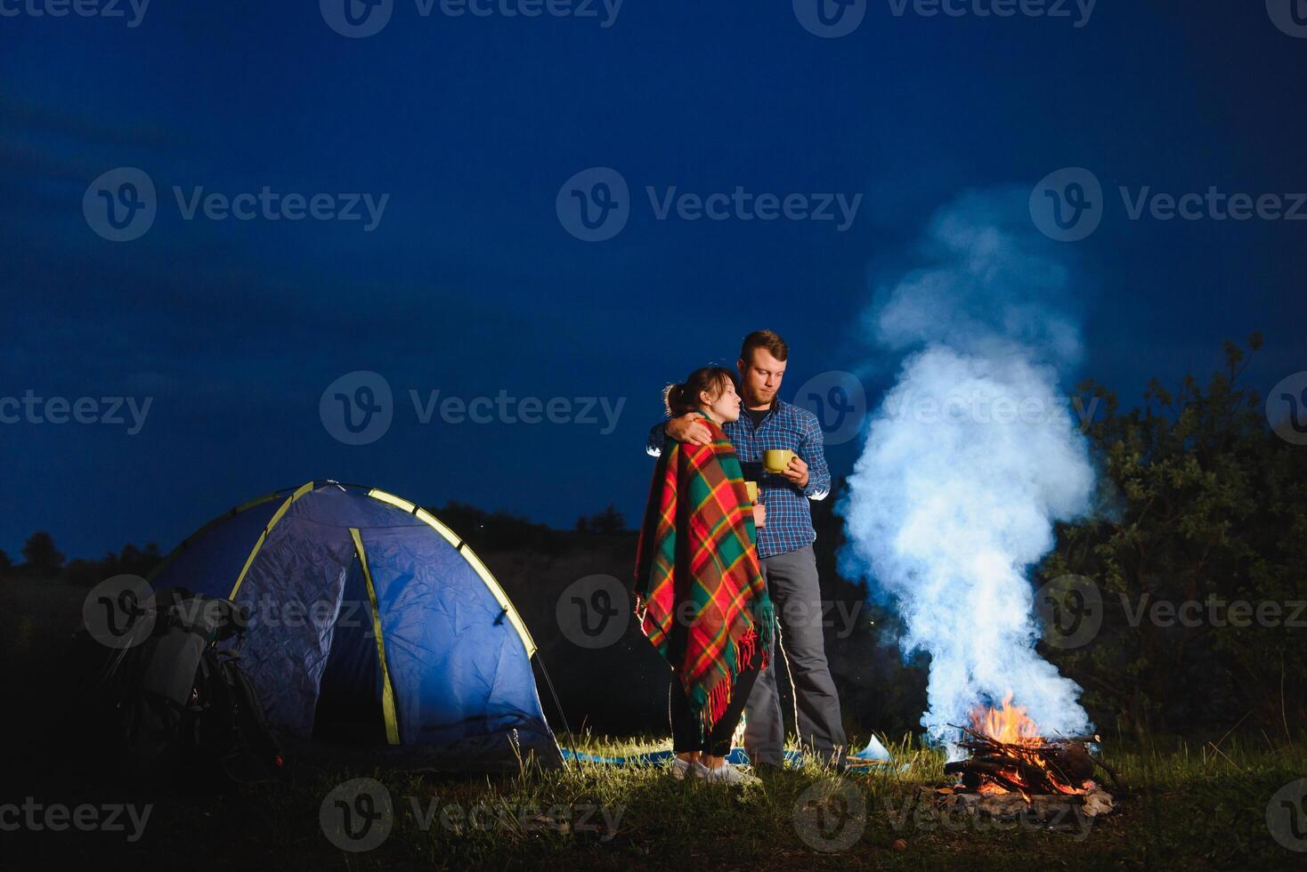 amoroso casal caminhantes desfrutando cada outro, em pé de fogueira às noite debaixo tarde céu perto árvores e barraca. romântico acampamento perto floresta dentro a montanhas foto