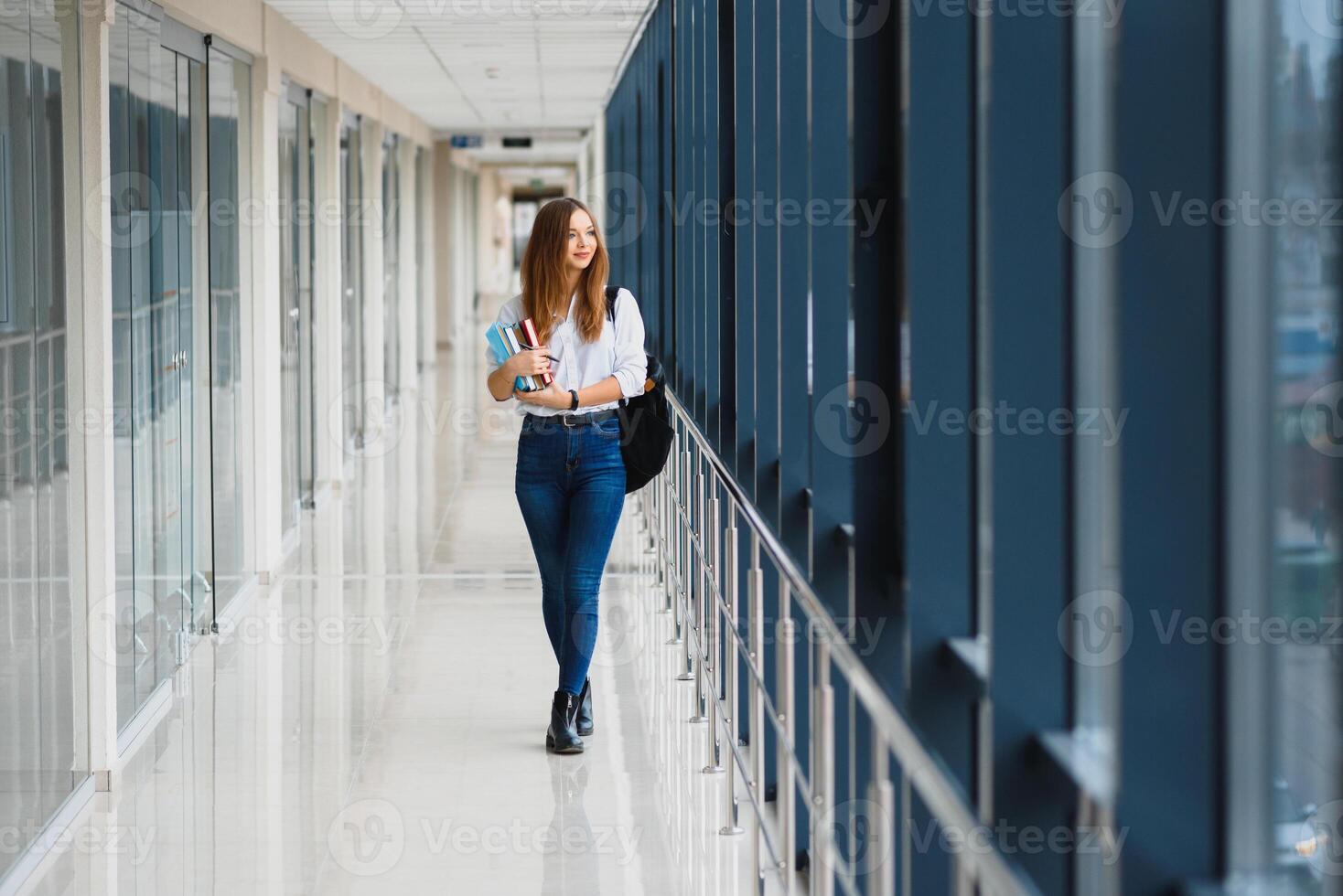 alegre morena aluna menina com Preto mochila detém livros dentro moderno prédio. fêmea aluna em pé com livros dentro Faculdade corredor foto