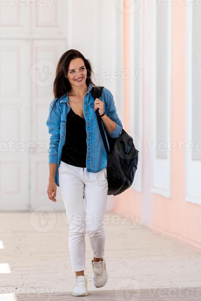 jovem mulher com grandes Castanho cabelo caminhando com mochila. frente visualizar. branco jeans e azul camisa. sorridente beleza. luz corredor fundo. foto